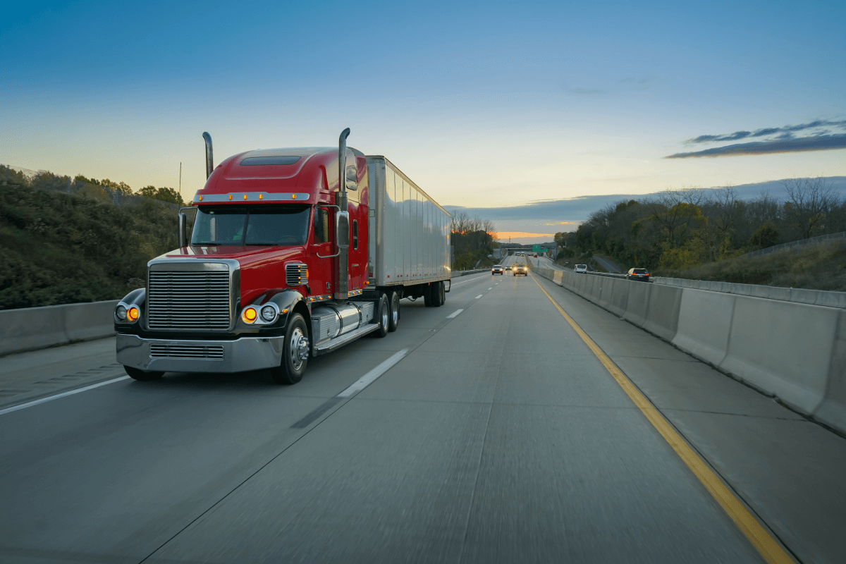 Bright red American modern long haul big rig semi truck with dry van semi trailer transporting commercial cargo moving on the wide multiline green highway road